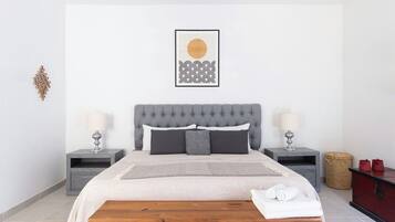 Un dormitorio con cabecera tapizada gris, dos mesitas de noche con lámparas, una cama con colcha blanca y gris estampada, y un banco de madera con toallas.