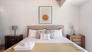 Una cama bien hecha con un tapete color beige, dos mesitas de noche con lámparas y una obra enmarcada sobre la cama.