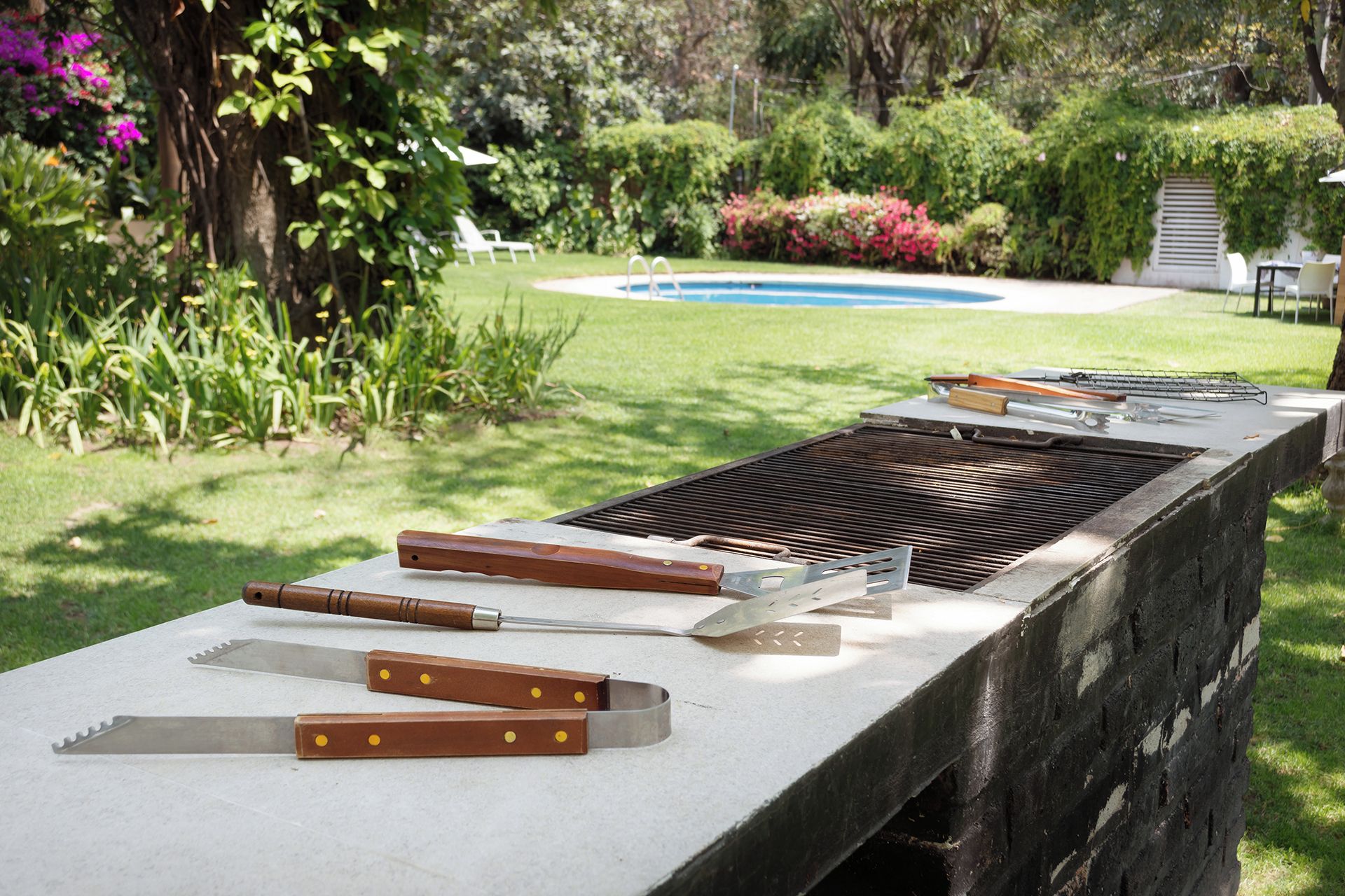 Una parrilla con diversos utensilios y una piscina al fondo.
