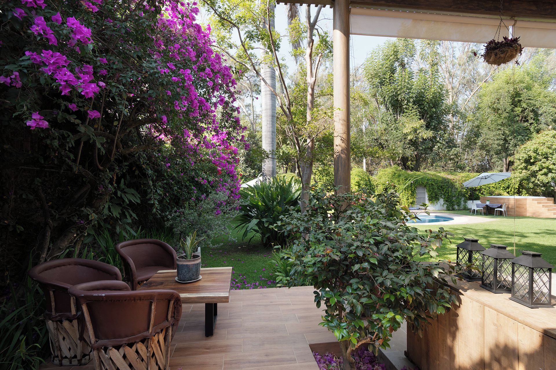A covered patio with a table and chairs, surrounded by lush greenery and flowering plants.