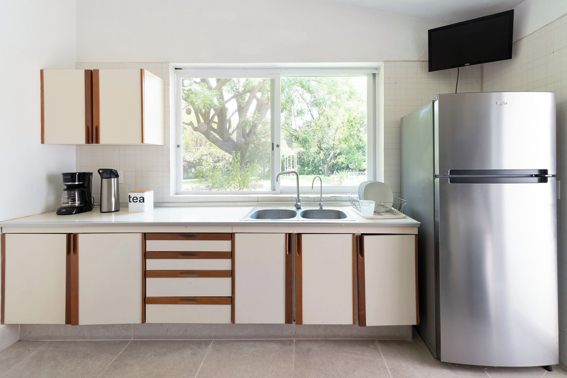 A modern kitchen with a stainless steel refrigerator, double sink, and a window with a view of greenery.