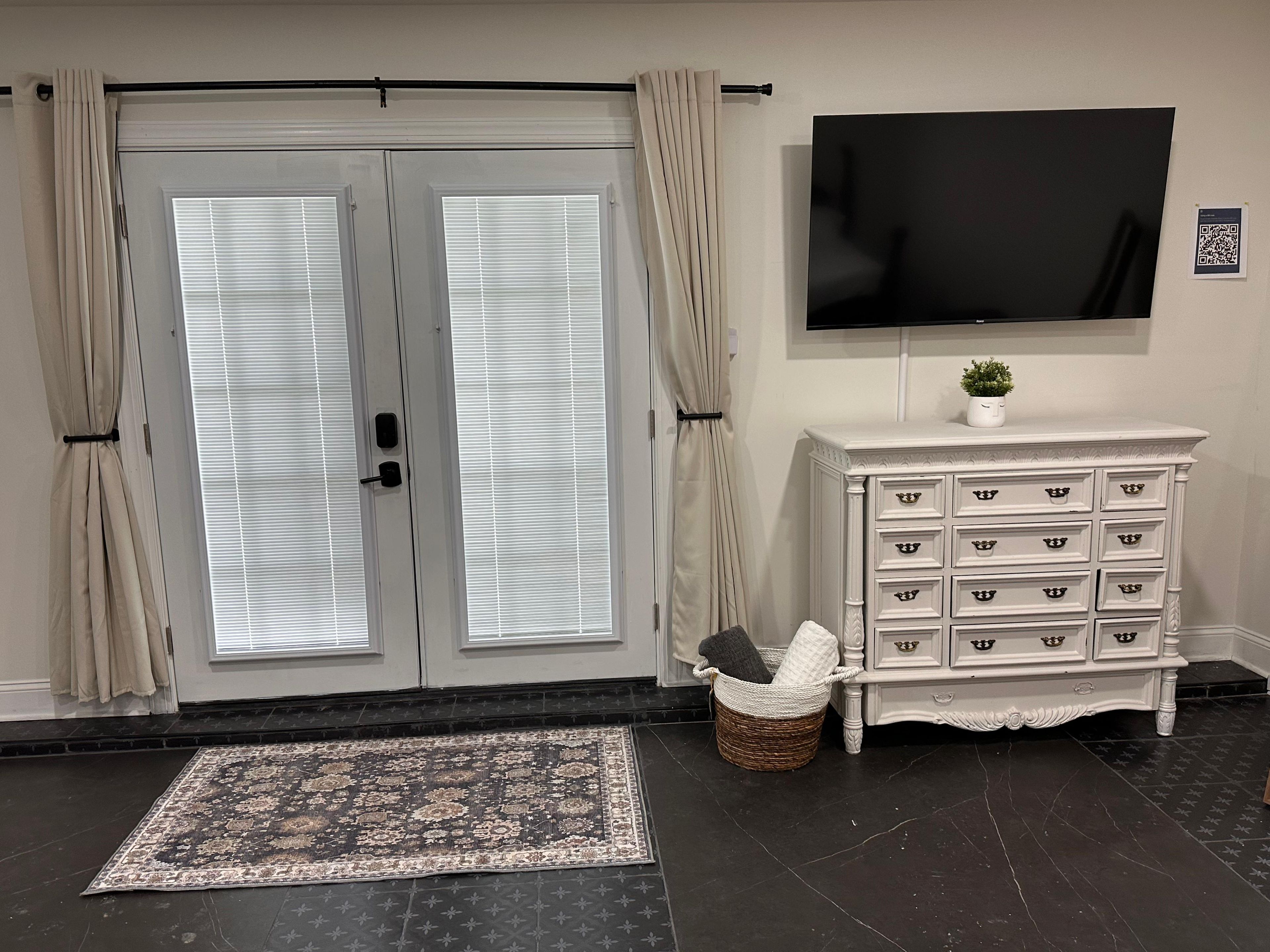 A room with a large double door, a flat-screen TV mounted on the wall, a white dresser with multiple drawers, and a woven basket on the floor.