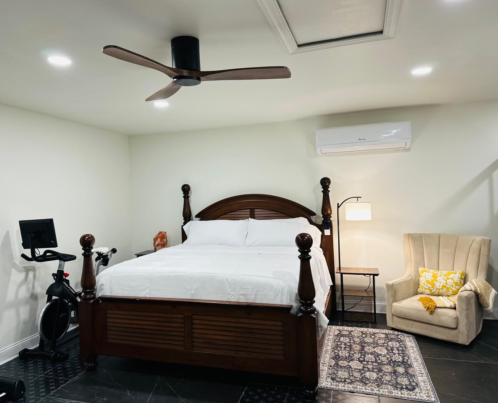 A bedroom with a wooden four-poster bed, a ceiling fan, a treadmill, a chair with a cushion, and a patterned rug.