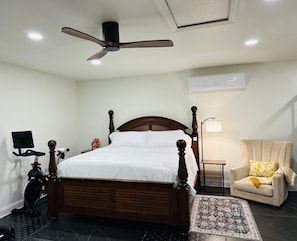 Un dormitorio con una cama con dosel de madera, ventilador de techo, cinta de correr, una silla con cojín y una alfombra estampada.