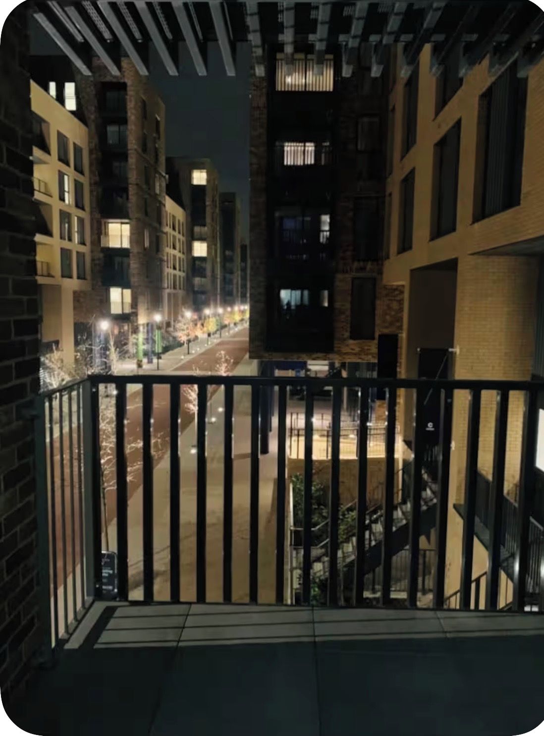 Een nachtzicht vanaf een balkon met een metalen hekwerk, uitkijkend over een straat met gebouwen en een heldere hemel.