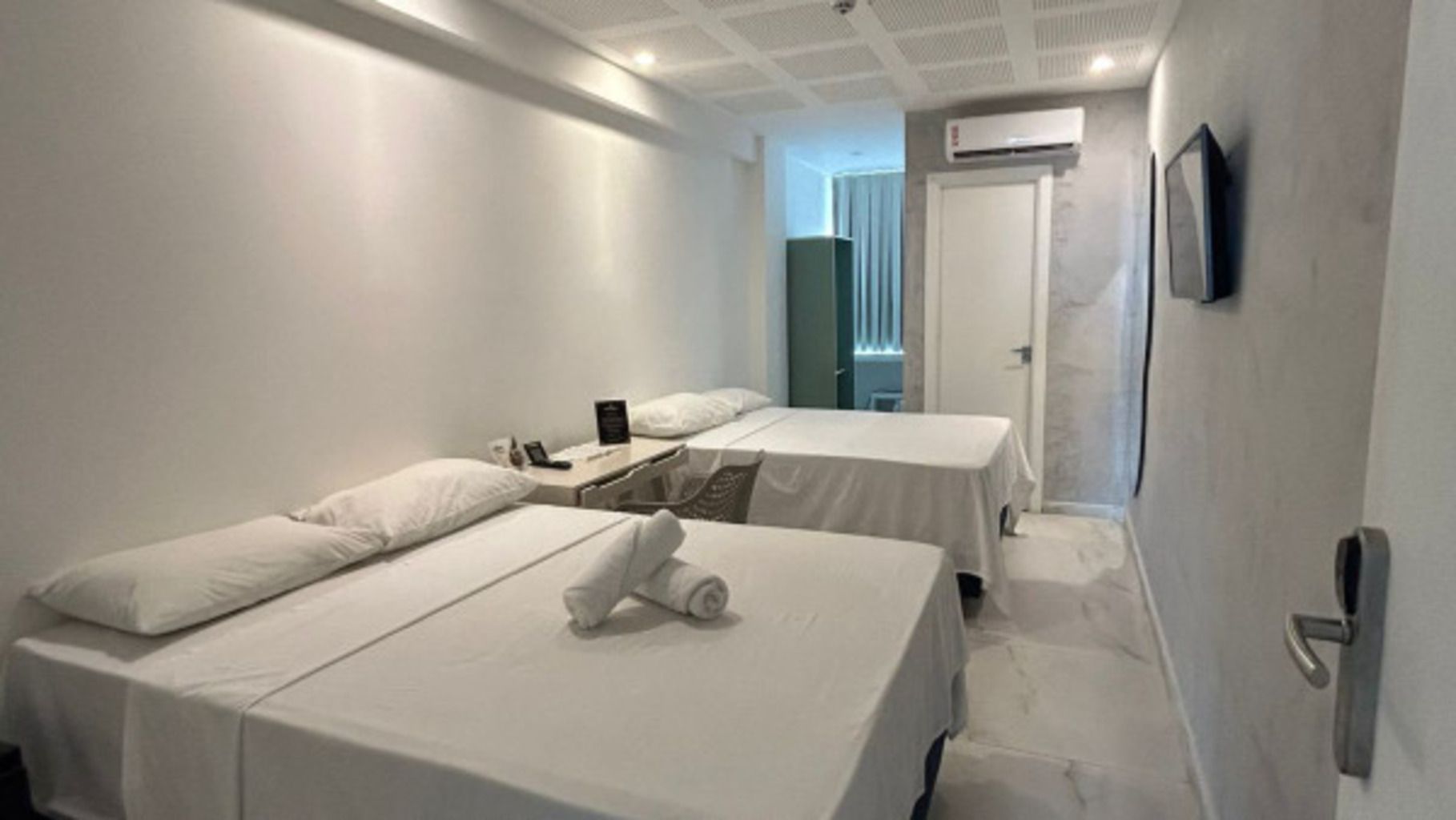 Quarto de hotel com duas camas, uma mesinha de cabeceira, TV na parede e aparelho de ar condicionado.