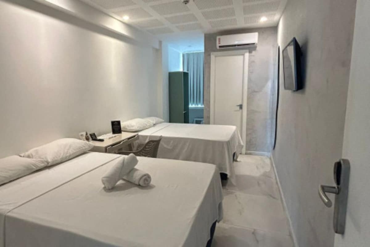 A hotel room with two beds, a bedside table, a wall-mounted TV, and an air conditioning unit.