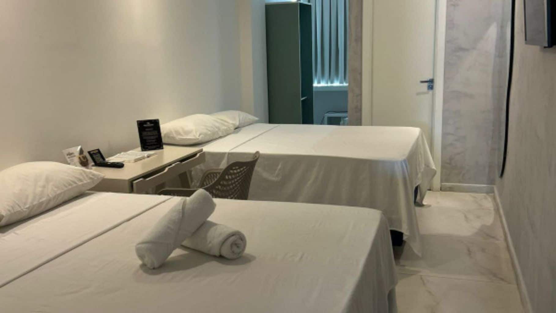 A hotel room with two beds, a desk with a phone and a sign, and a window with blinds.