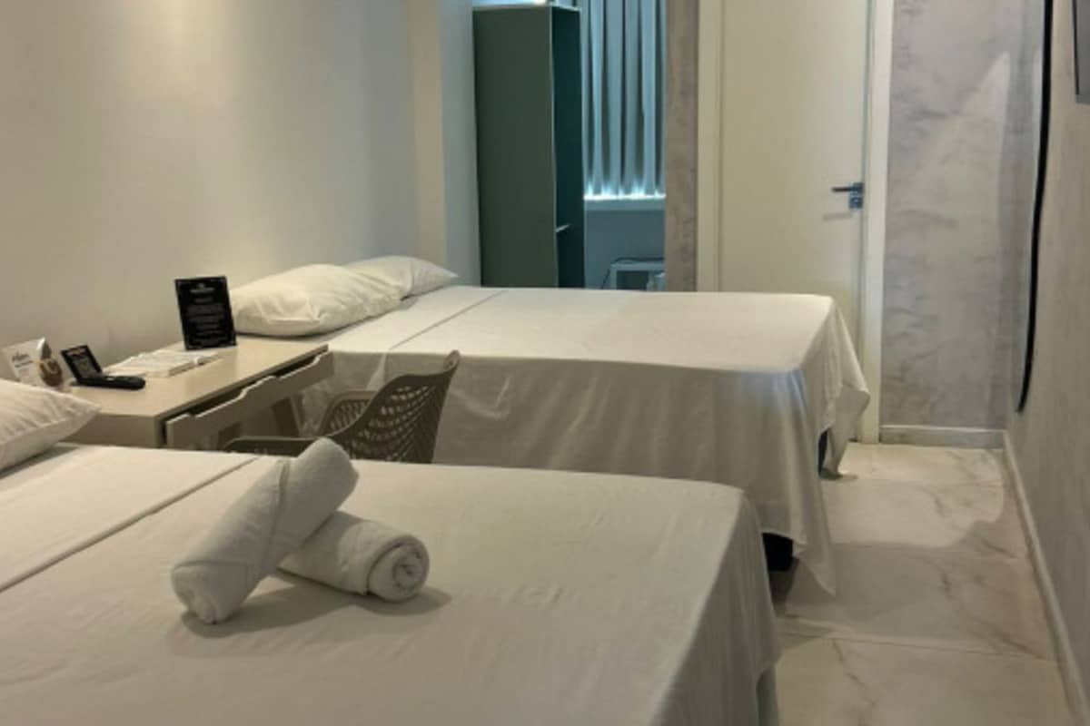 A hotel room with two beds, a desk with a phone and a sign, and a window with blinds.