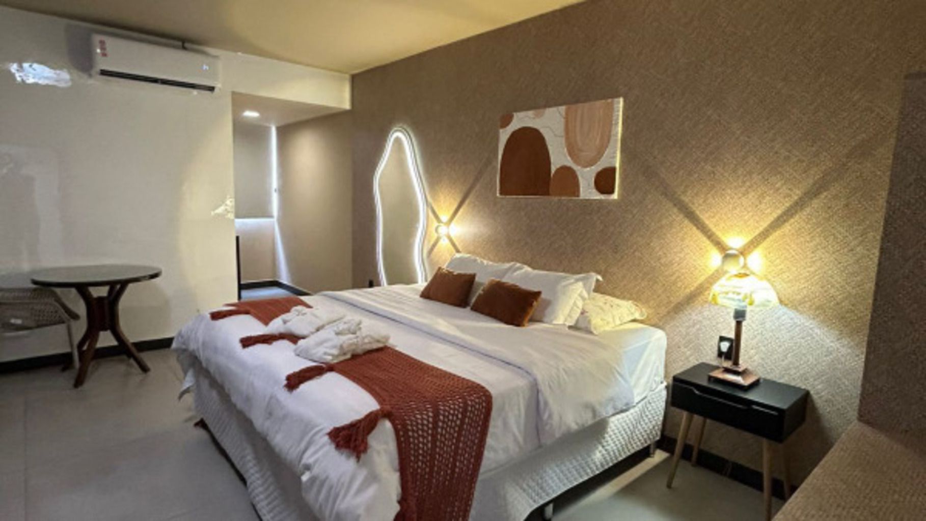 Quarto de hotel com cama, mesa de cabeceira e luminária fixada na parede.