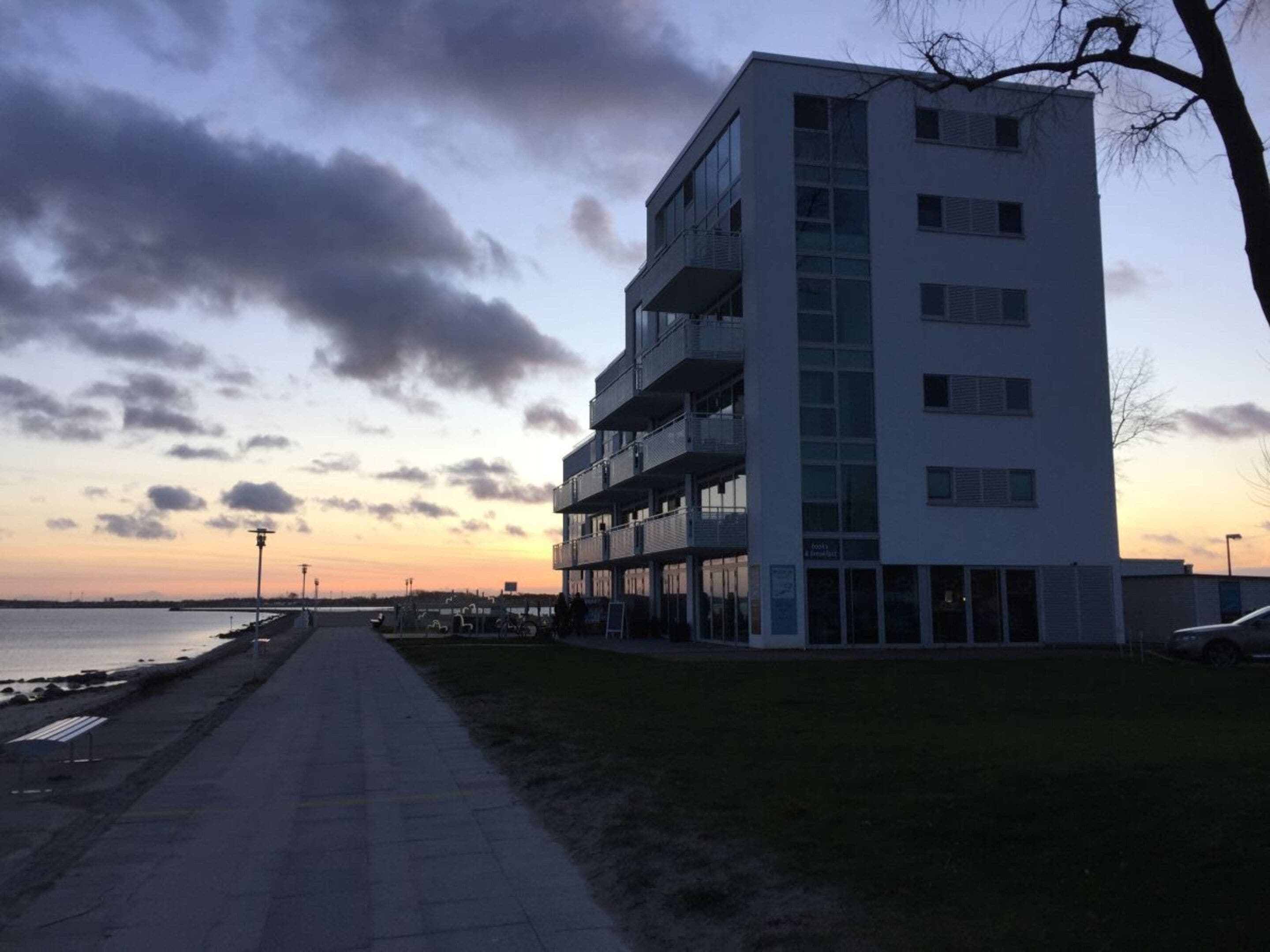 Un edificio de varios pisos con balcones, un camino empedrado y un cuerpo de agua al fondo.