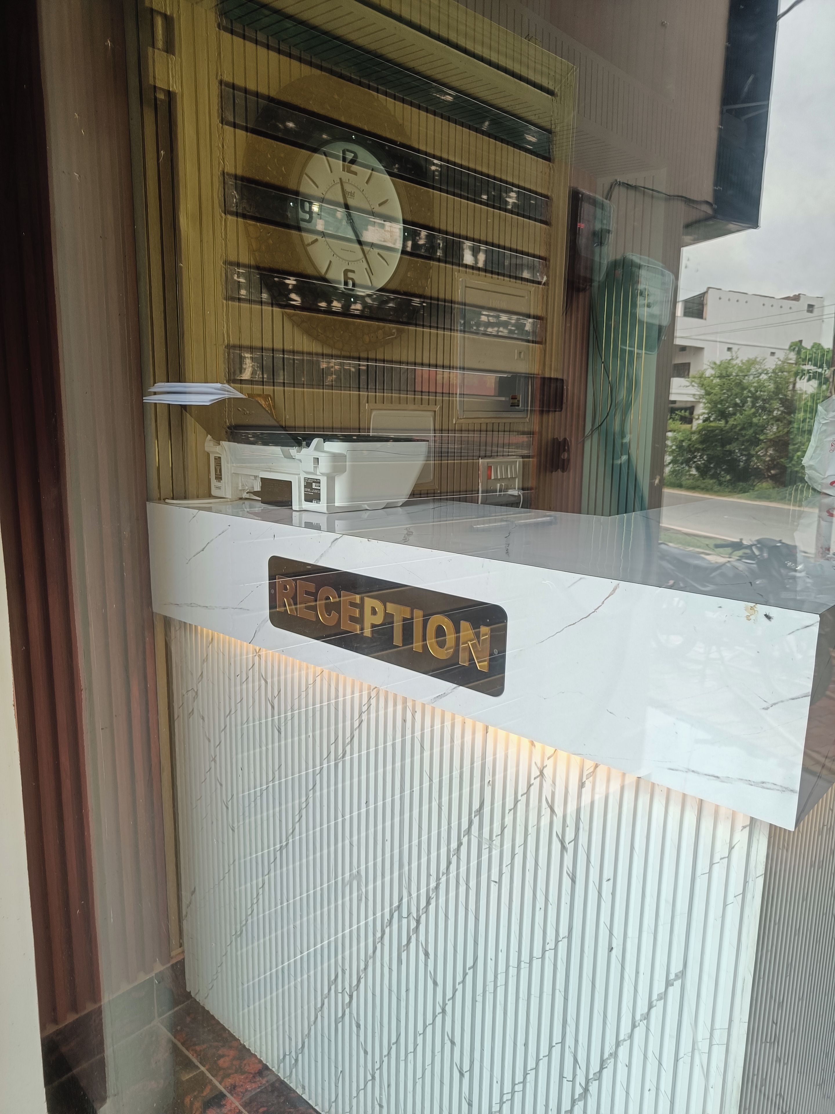 A reception desk with a clock above it, a sign that reads 'RECEPTION', and a window with a view of buildings and greenery.
