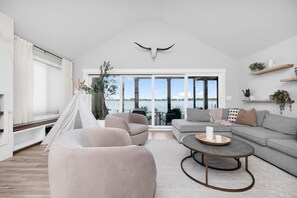 Ein geräumiges Wohnzimmer mit großem Fenster, weißer Decke und Blick auf das Wasser.