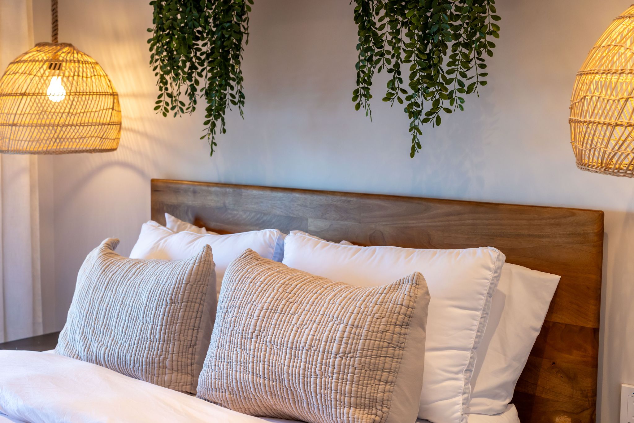 A bed with white bedding and textured pillows, a wooden headboard, and hanging plants.