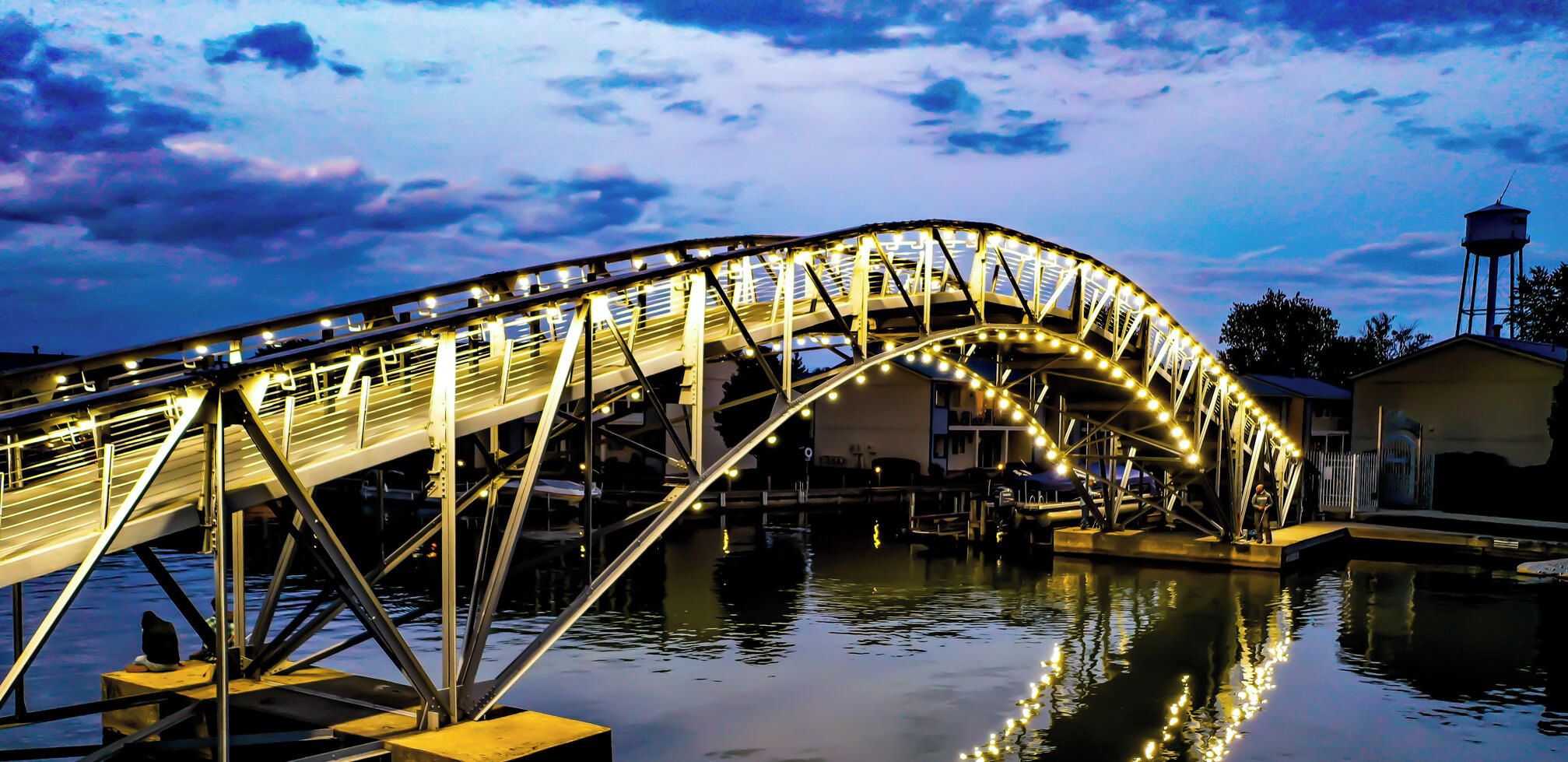 Un pont en acier illuminé, qui enjambe une étendue d’eau, avec un château d’eau en arrière-plan.