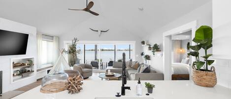A modern living room with a large TV, a ceiling fan, and a view of the water.