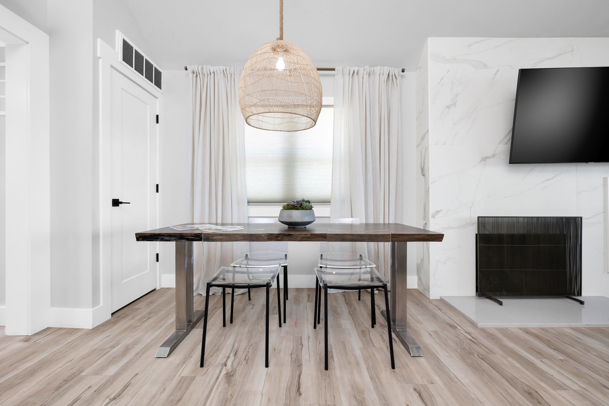 A modern dining area with a wooden table, two chairs, a hanging light fixture, a TV mounted on the wall, and a fireplace.