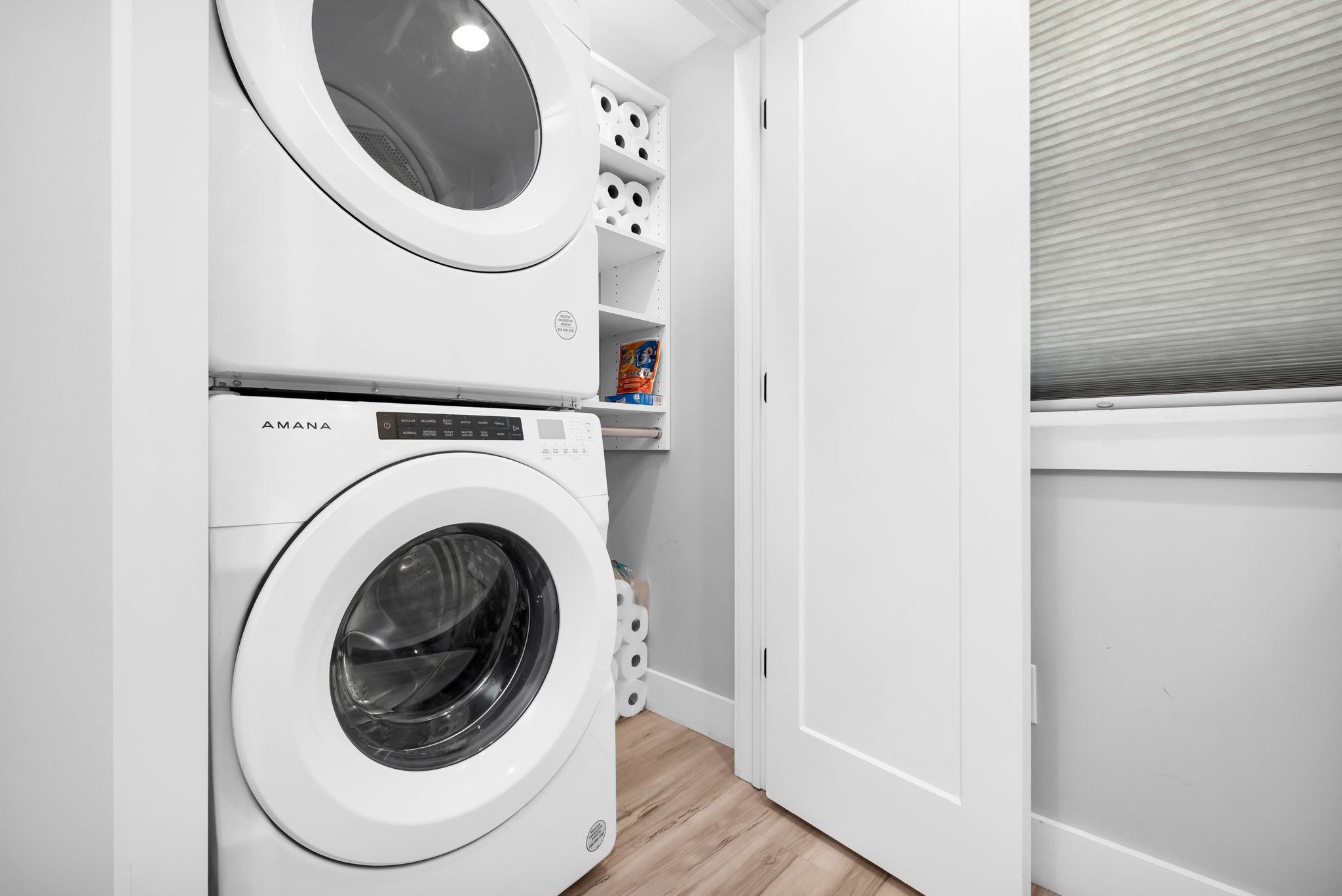 A compact laundry area with two white washing machines, a shelf with laundry supplies, and a door leading to another room.