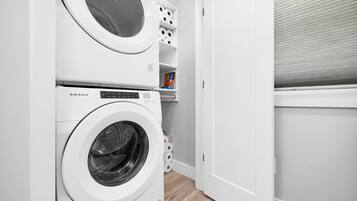 A compact laundry area with two white washing machines, a shelf with laundry supplies, and a door leading to another room.
