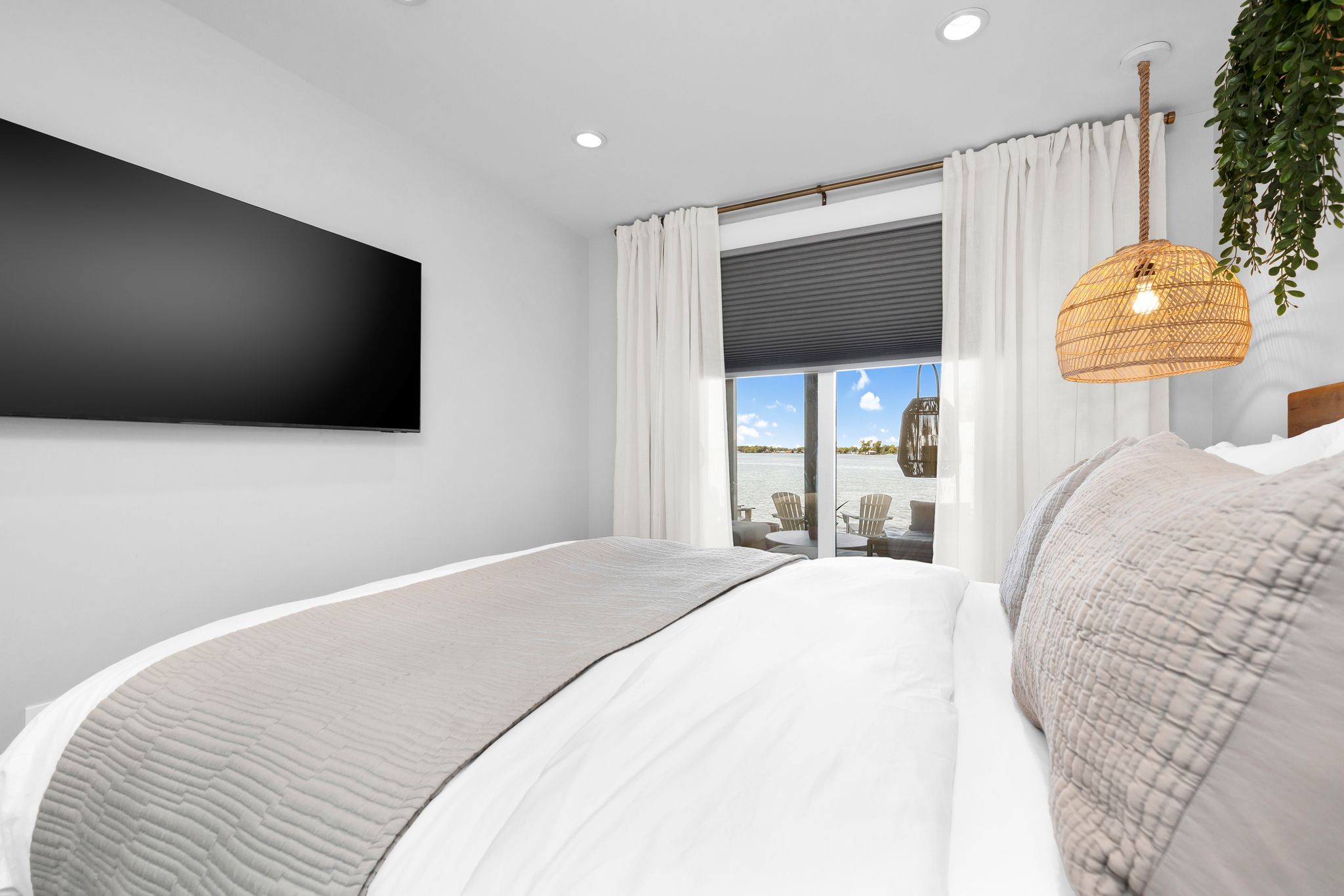 A modern bedroom with a large bed, a flat-screen TV, a wicker hanging light fixture, and a view of the outdoors.