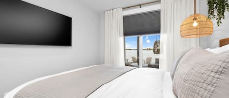 A modern bedroom with a large bed, a flat-screen TV, a wicker hanging light fixture, and a view of the outdoors.