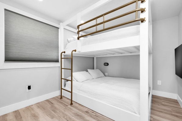 A bunk bed room with a ladder, a window with blinds, and a flat-screen TV on the wall.