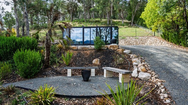 Un jardin avec un banc en pierre, une poubelle et une structure en verre offrant une vue sur le paysage.