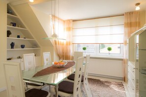 A dining area with a glass table, white chairs, a bowl of fruit, and a view of the outside through a window with curtains. - Regina House 11 (Timmendorfer Strand)