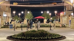 A nighttime view of the Boardwalk entrance with illuminated signage, a landscaped area with plants, and people walking around. - Comfort Meets Space - Feel Right at Home (Cypress)