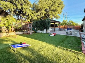 Un patio trasero con un columpio, un parque infantil, una mesa de picnic y una cancha de bochás.