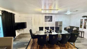 A modern dining area with a glass table, black upholstered chairs, a chandelier, a flat-screen TV, and a white door.