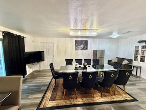 A modern dining area with a glass table, black upholstered chairs, a chandelier, a flat-screen TV, and a white door.