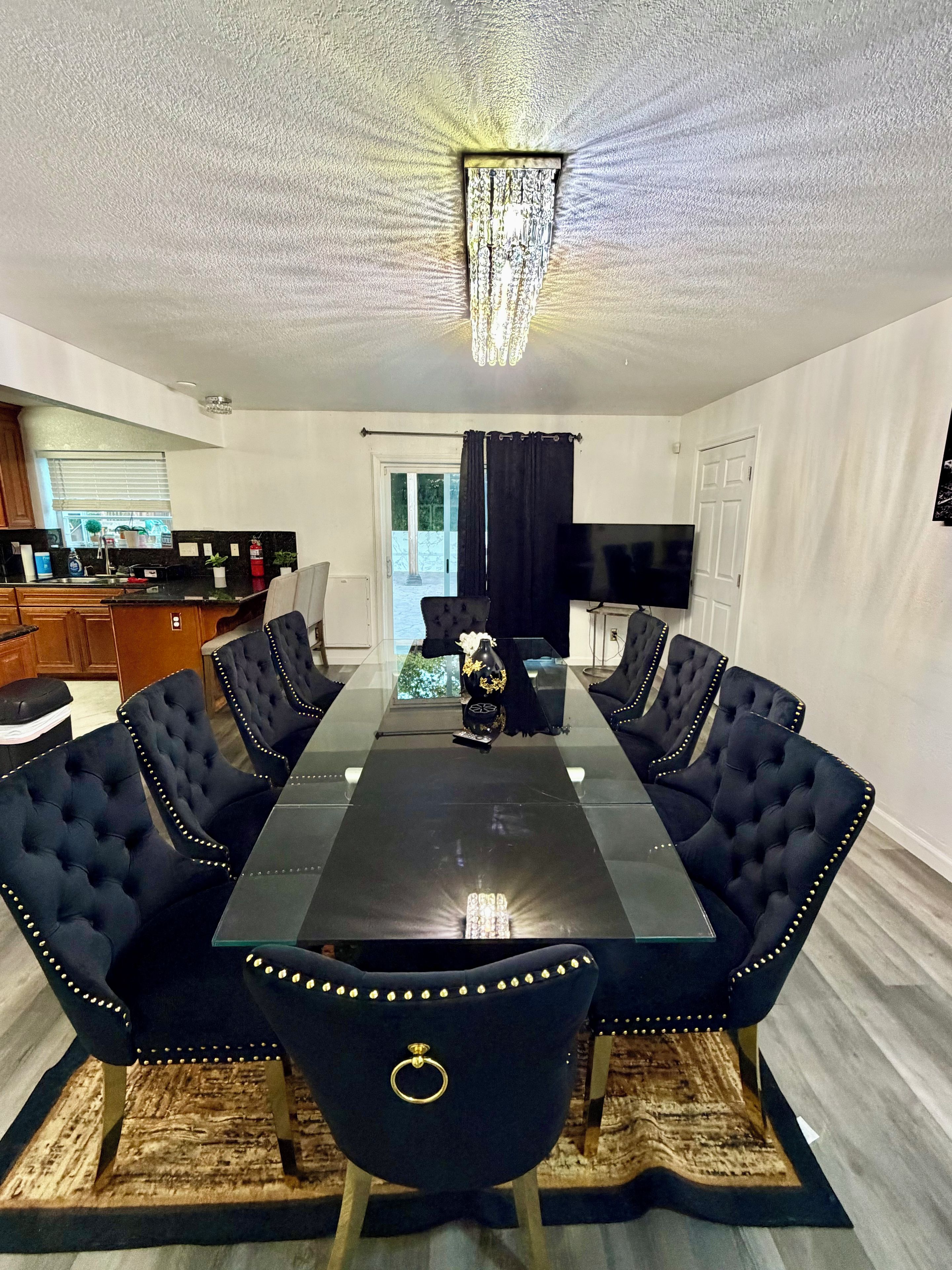A dining area with a glass-top table, black upholstered chairs, a chandelier, and a kitchen in the background.