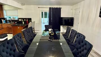 A dining area with a glass-top table, black upholstered chairs, a chandelier, and a kitchen in the background.