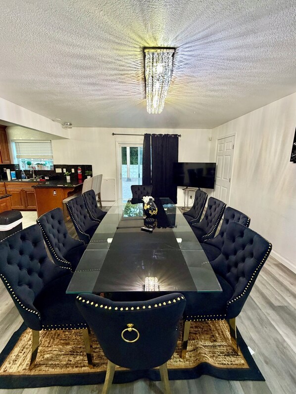 A dining area with a glass-top table, black upholstered chairs, a chandelier, and a kitchen in the background.