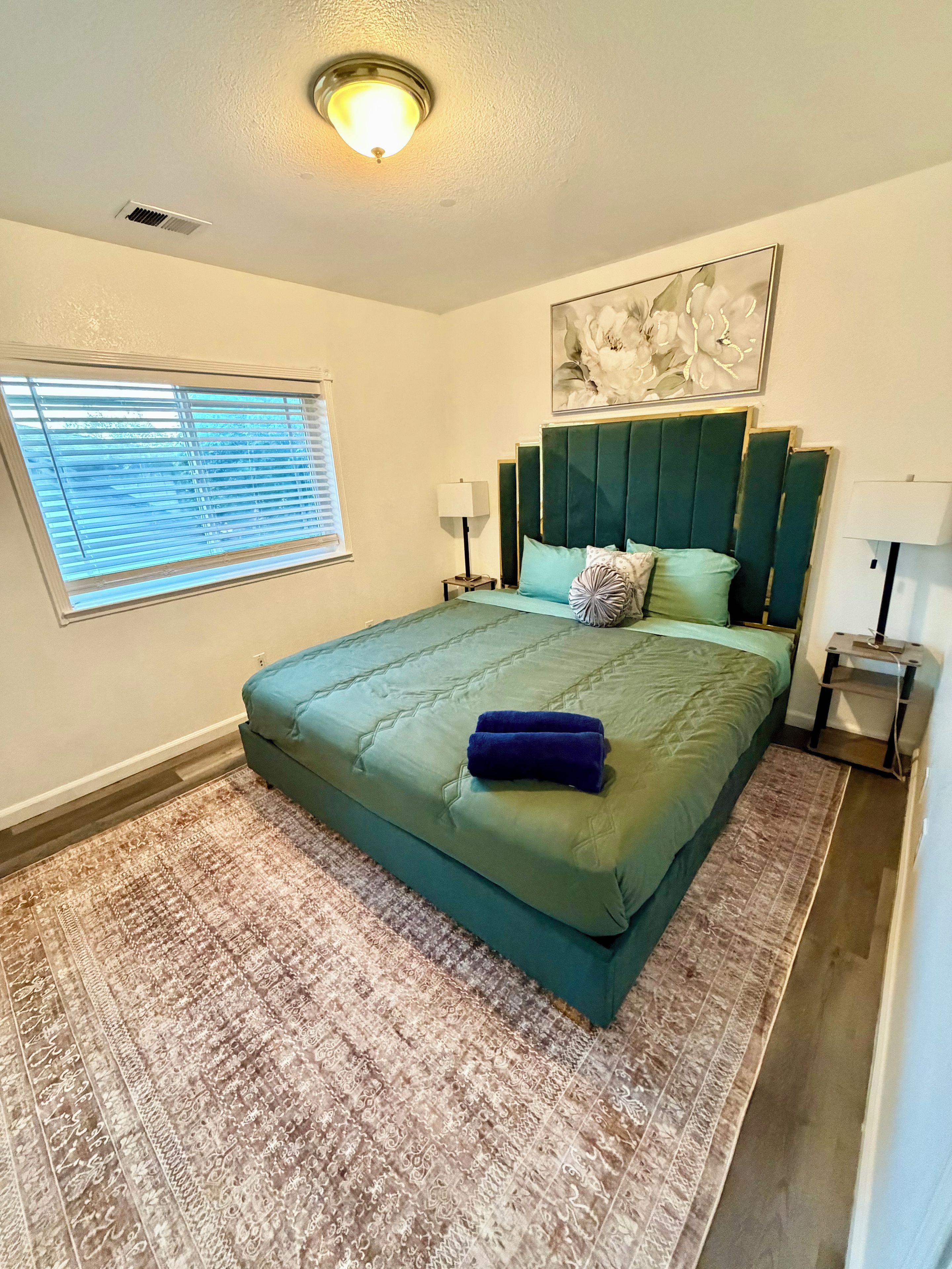 A bedroom with a bed, a window with blinds, a ceiling light, and a wall-mounted artwork.