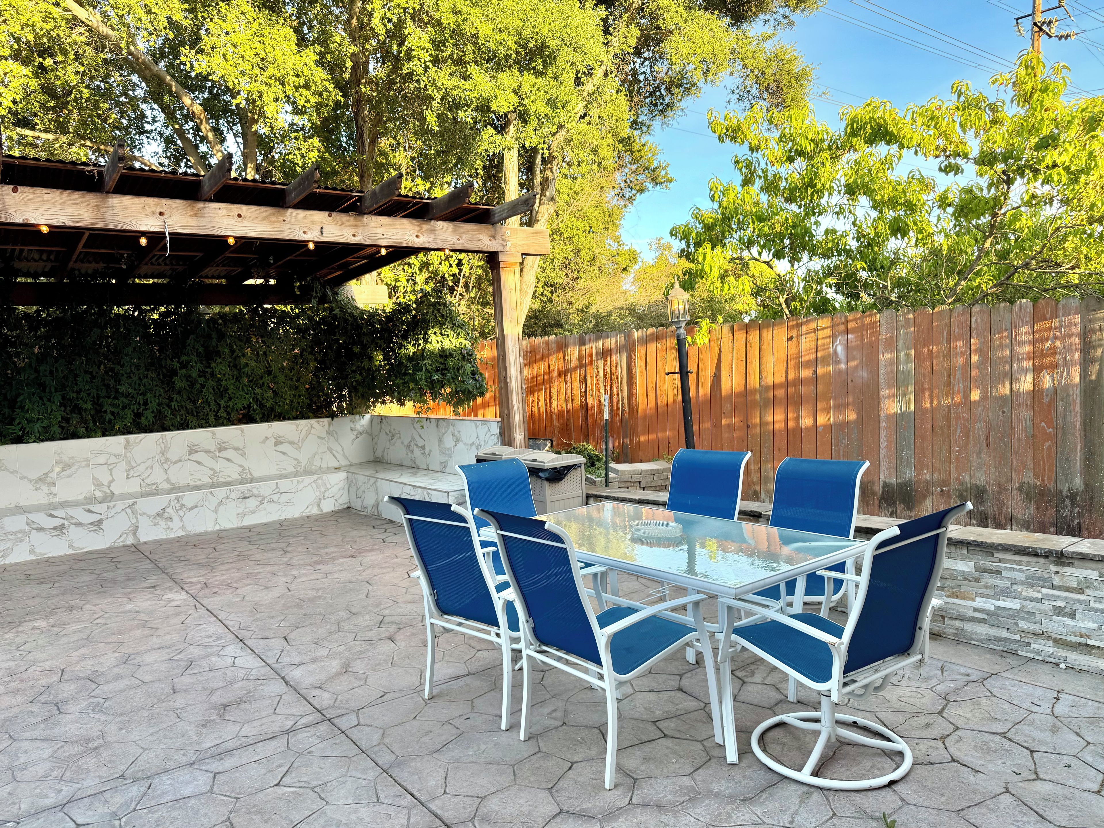 Une terrasse extérieure avec une table en verre et des chaises bleues, entourée d’une pergola en bois et d’une clôture en bois.