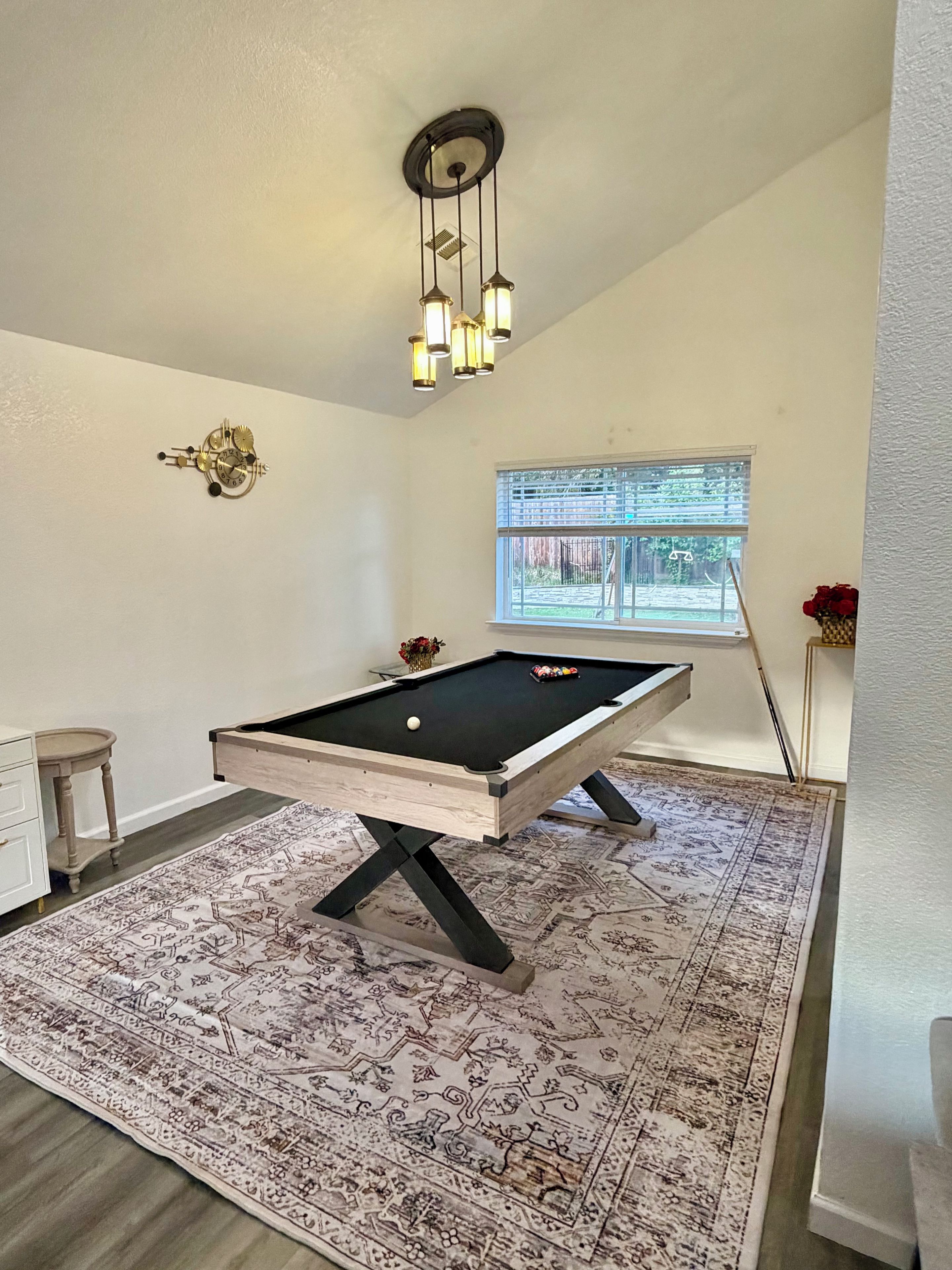 A pool table with a patterned rug, a clock on the wall, and a window with blinds.
