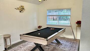 A pool table with a patterned rug, a clock on the wall, and a window with blinds.
