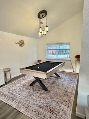 A pool table with a patterned rug, a clock on the wall, and a window with blinds.