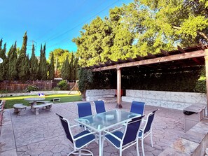 Un patio al aire libre con una mesa y sillas de vidrio, rodeado por un patio de piedra y una pérgola de madera.