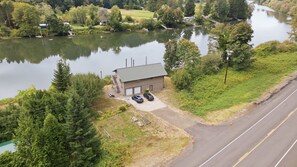 Ein kleines Gebäude mit Garage und zwei geparkten Autos vor der Fassade, neben einem Fluss mit einer Brücke im Hintergrund.