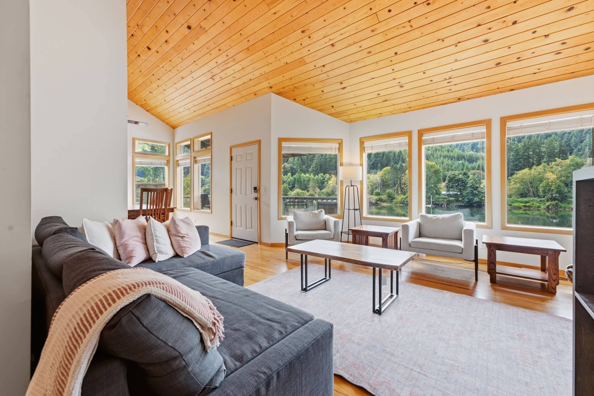 A spacious living room with a high ceiling, large windows offering a view of a forest, a grey sofa, a coffee table, and a TV.