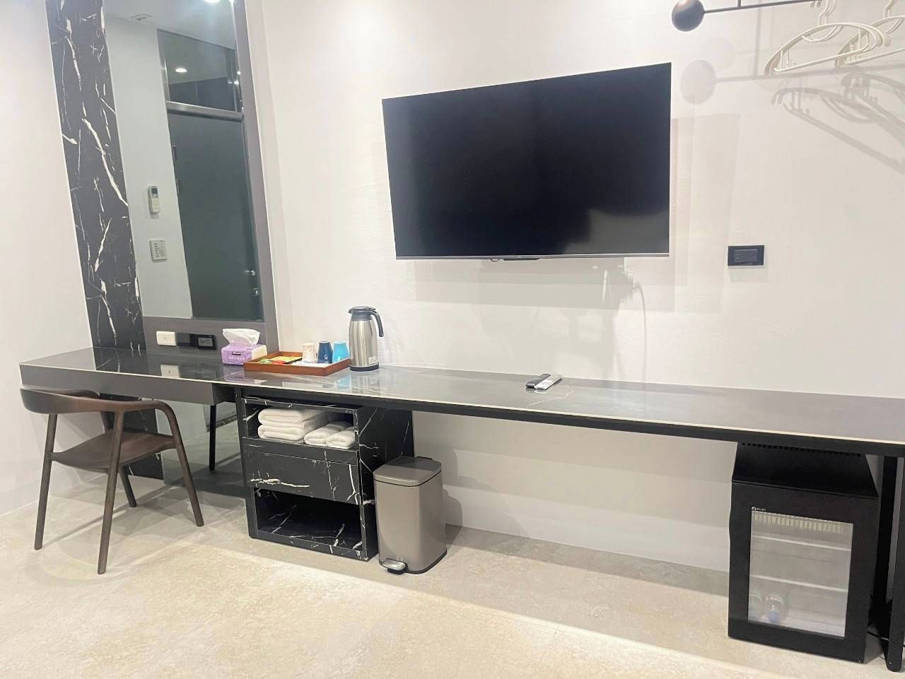 A modern hotel room with a long, dark-colored desk, a flat-screen TV mounted on the wall, a small trash bin, and a mirror.