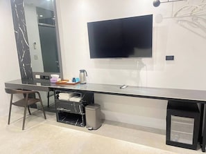 Habitación de hotel moderna con un escritorio largo y oscuro, un televisor de pantalla plana montado en la pared, un cubo de basura pequeño y un espejo.