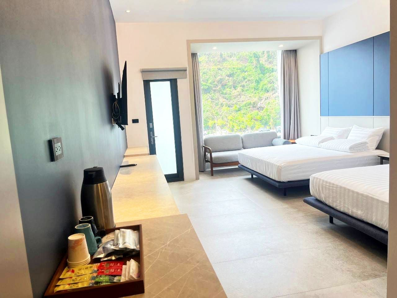 A hotel room with two beds, a sofa, a large window with curtains, a flat-screen TV mounted on the wall, and a tray table with various items.