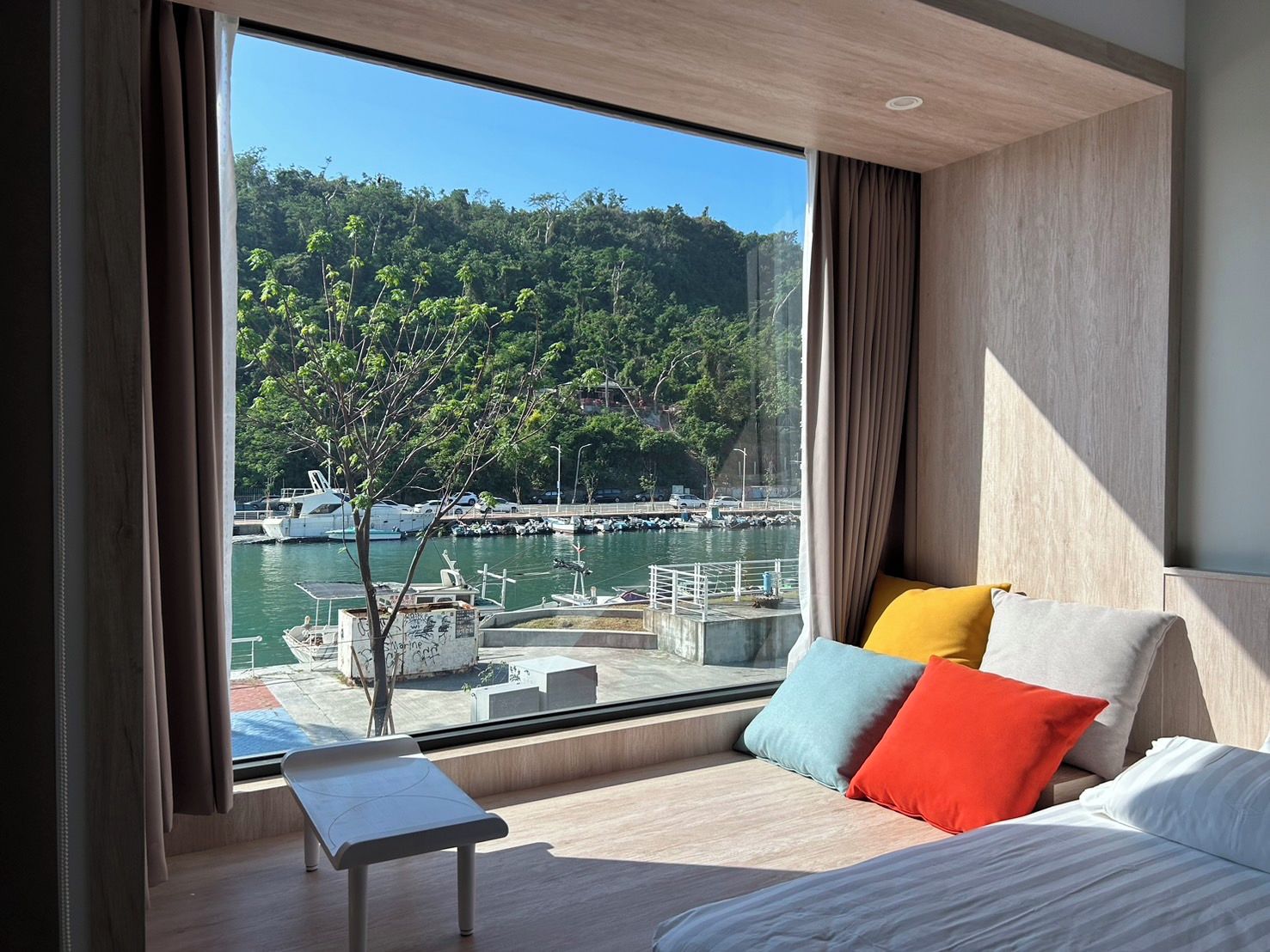 Habitación de hotel moderna con un amplio ventanal que ofrece vistas a un puerto deportivo y colinas boscosas.