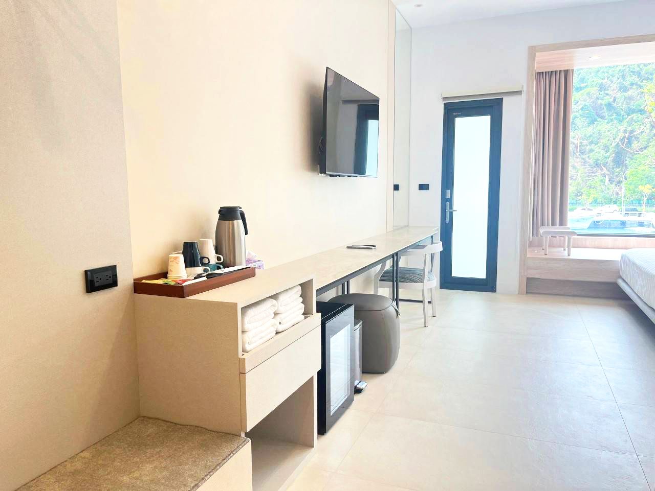 A modern hotel room with a flat-screen TV mounted on the wall, a small desk with a coffee maker, a trash bin, and a bed with a view of the outdoors.
