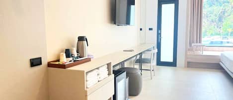 A modern hotel room with a flat-screen TV mounted on the wall, a small desk with a coffee maker, a trash bin, and a bed with a view of the outdoors.