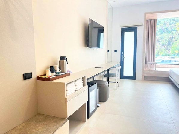 Una habitación de hotel moderna con televisión de pantalla plana montada en la pared, un escritorio pequeño con cafetera, un cesto de basura y una cama con vistas al exterior.